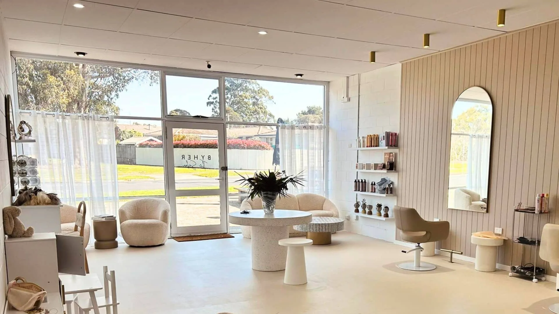 A modern, minimalist salon with light-coloured walls and floors, large windows with sheer curtains, a round table with a plant centrepiece, various chairs, shelves with hair products, and a tall wall mirror.